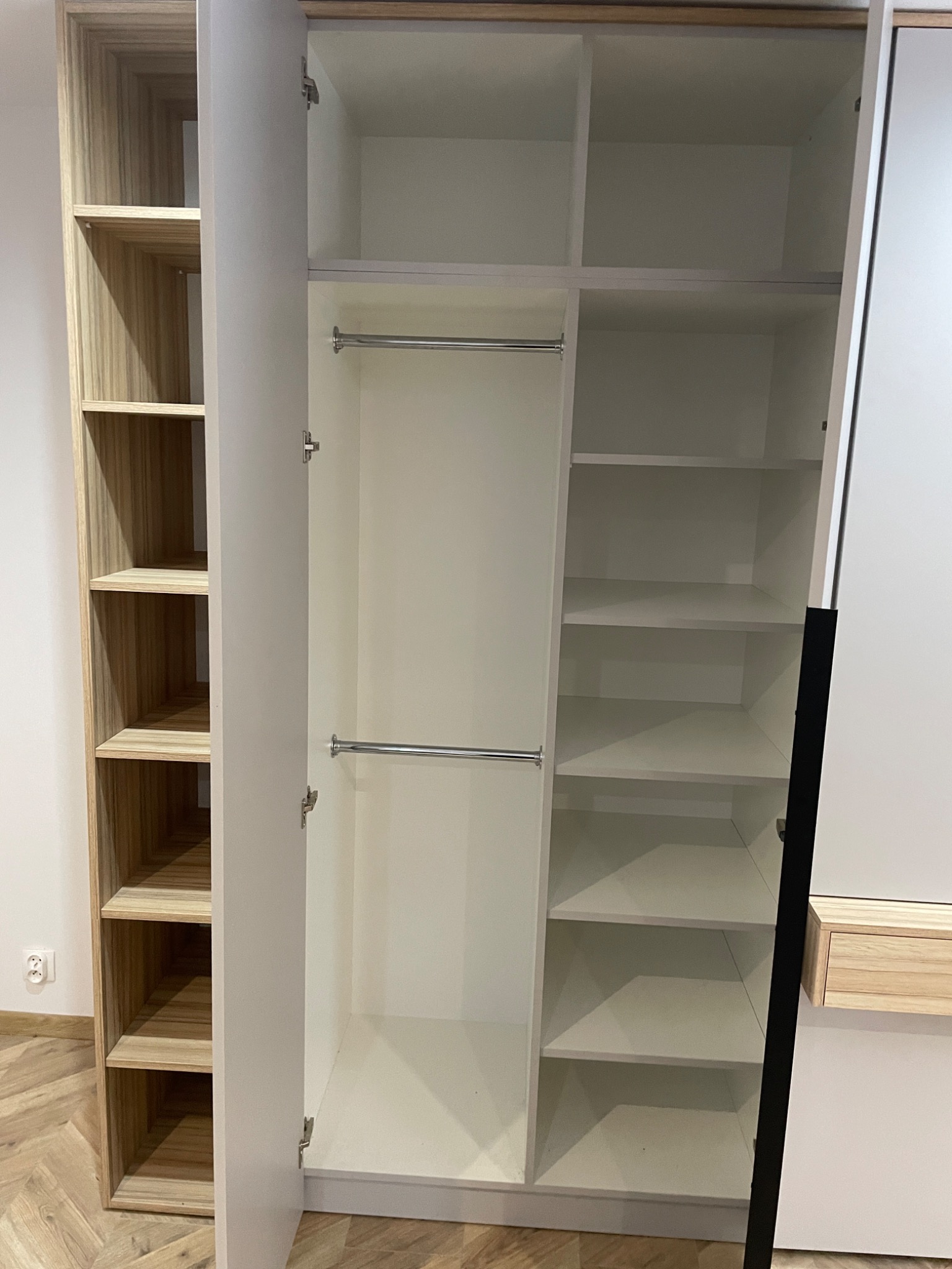 Otwarta szafa wnękowa z białymi półkami i drążkami na ubrania, obok regał z drewnianymi półkami, widoczna podłoga w jodełkę.