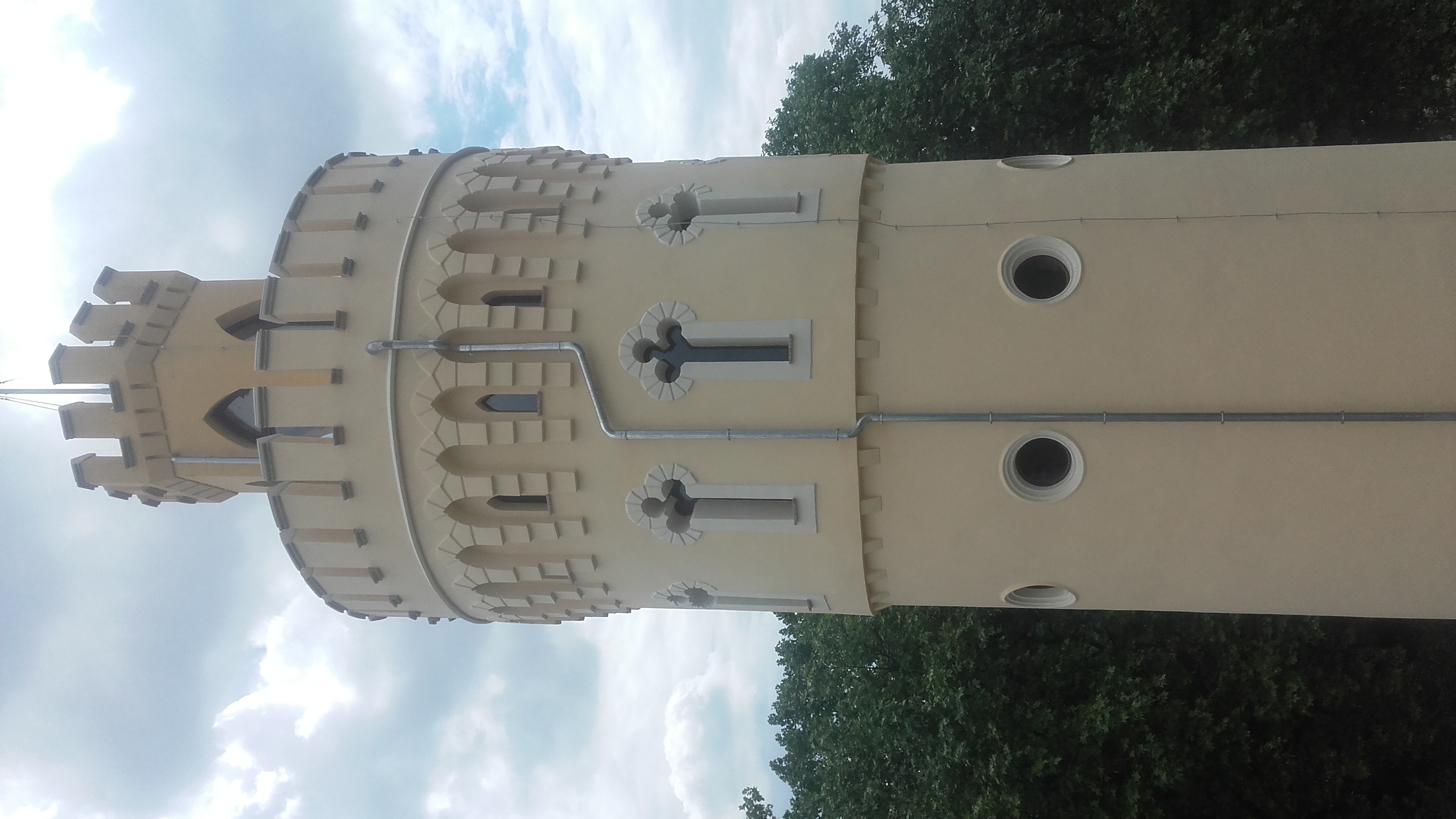 Wieża w stylu neogotyckim z kremową elewacją, ozdobnymi oknami i fragmentem rynny, widziana z dołu na tle drzew i pochmurnego nieba.