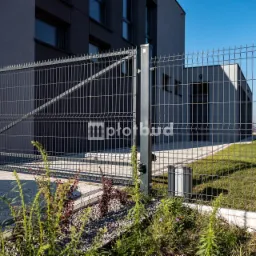 Nowoczesne ogrodzenie panelowe z bramą przesuwną, widoczne automatyczne sterowanie, szara kolorystyka, na tle budynku w minimalistycznym stylu, w tle komin przemysłowy.