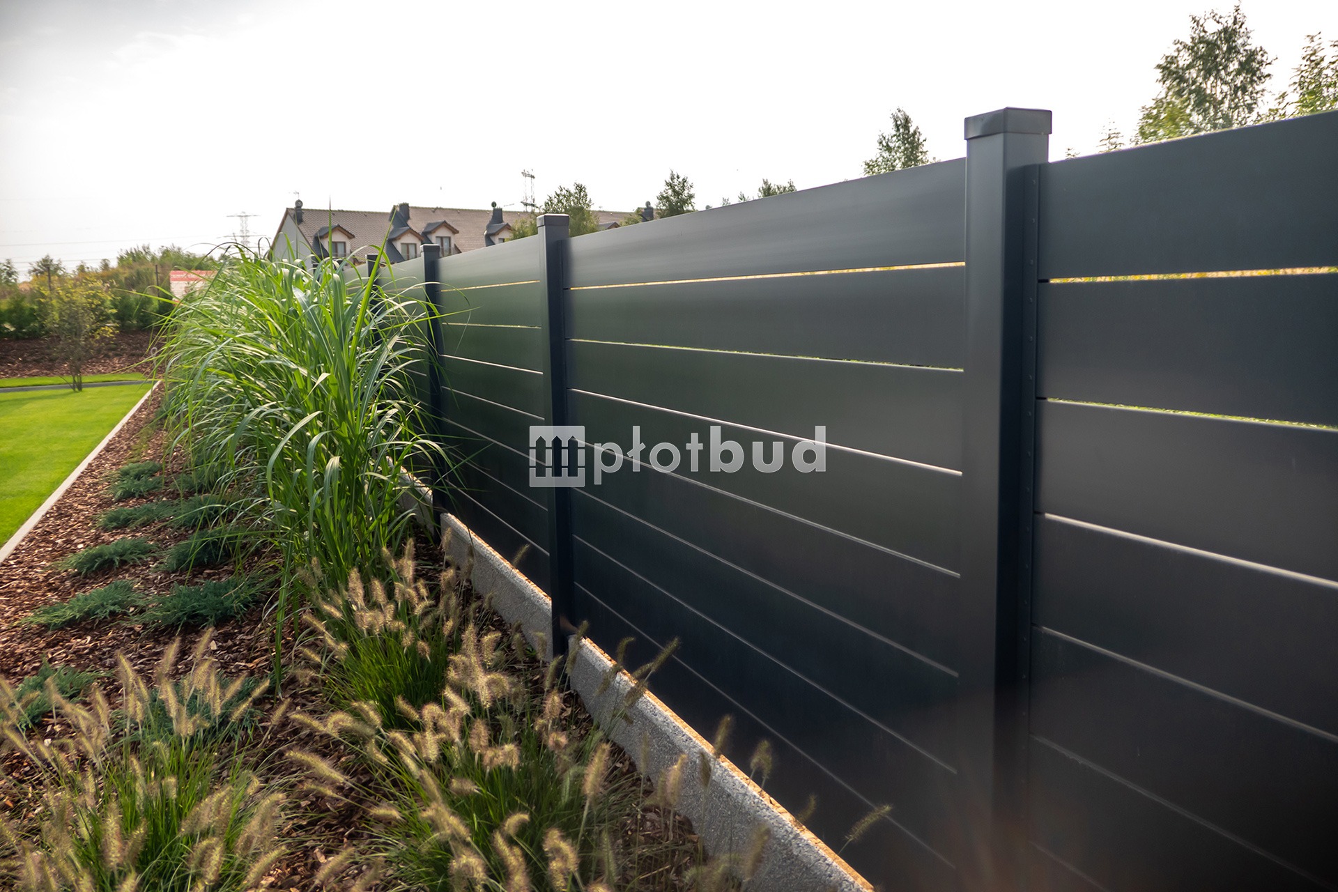 Nowoczesne, poziome, szare ogrodzenie z metalowych paneli, z dekoracyjnym obramowaniem z betonu architektonicznego i ozdobnymi trawami, widoczne logo firmy.