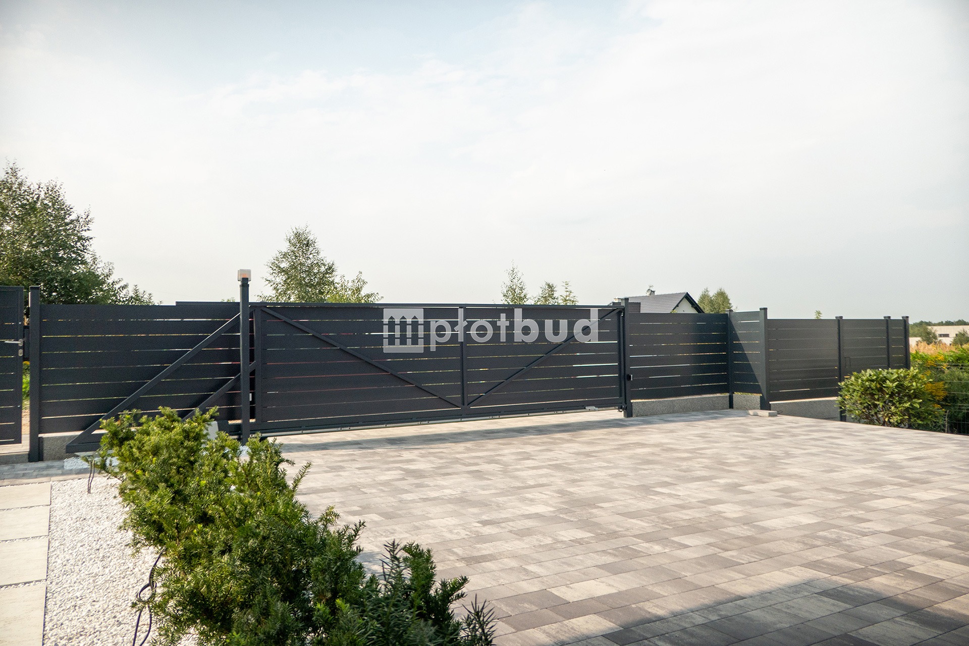 Nowoczesna, grafitowa brama przesuwna z poziomych profili, zintegrowana z ogrodzeniem panelowym, widoczny znak firmowy 'plotbud' na bramie, kostka brukowa na podjeździe.