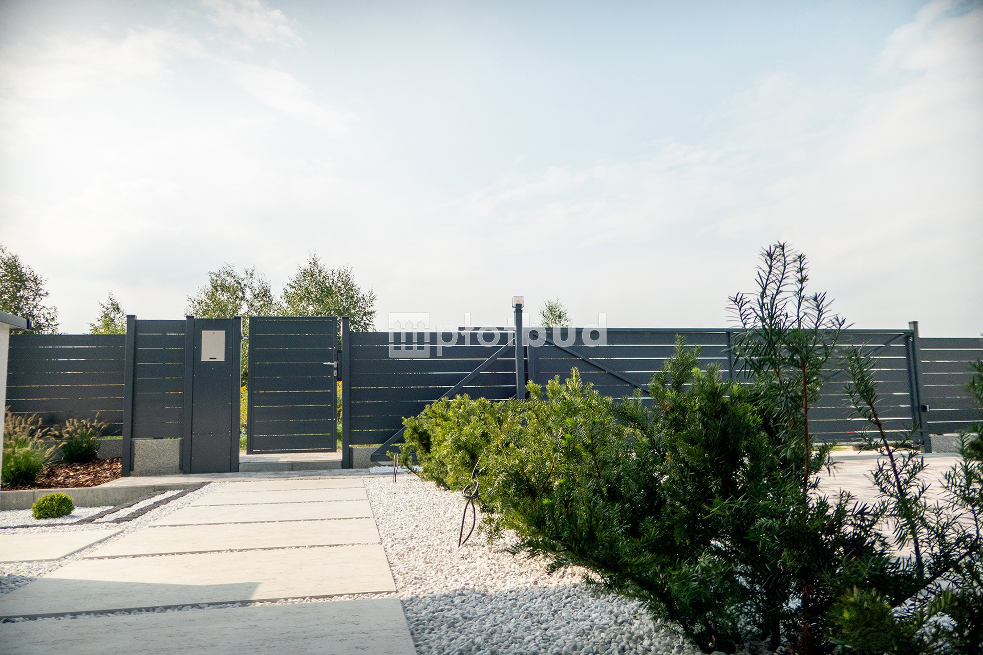 Nowoczesne ogrodzenie panelowe w Tarnowskich Górach z przesuwną bramą i furtką, widoczne zza ozdobnych krzewów i białego żwiru.