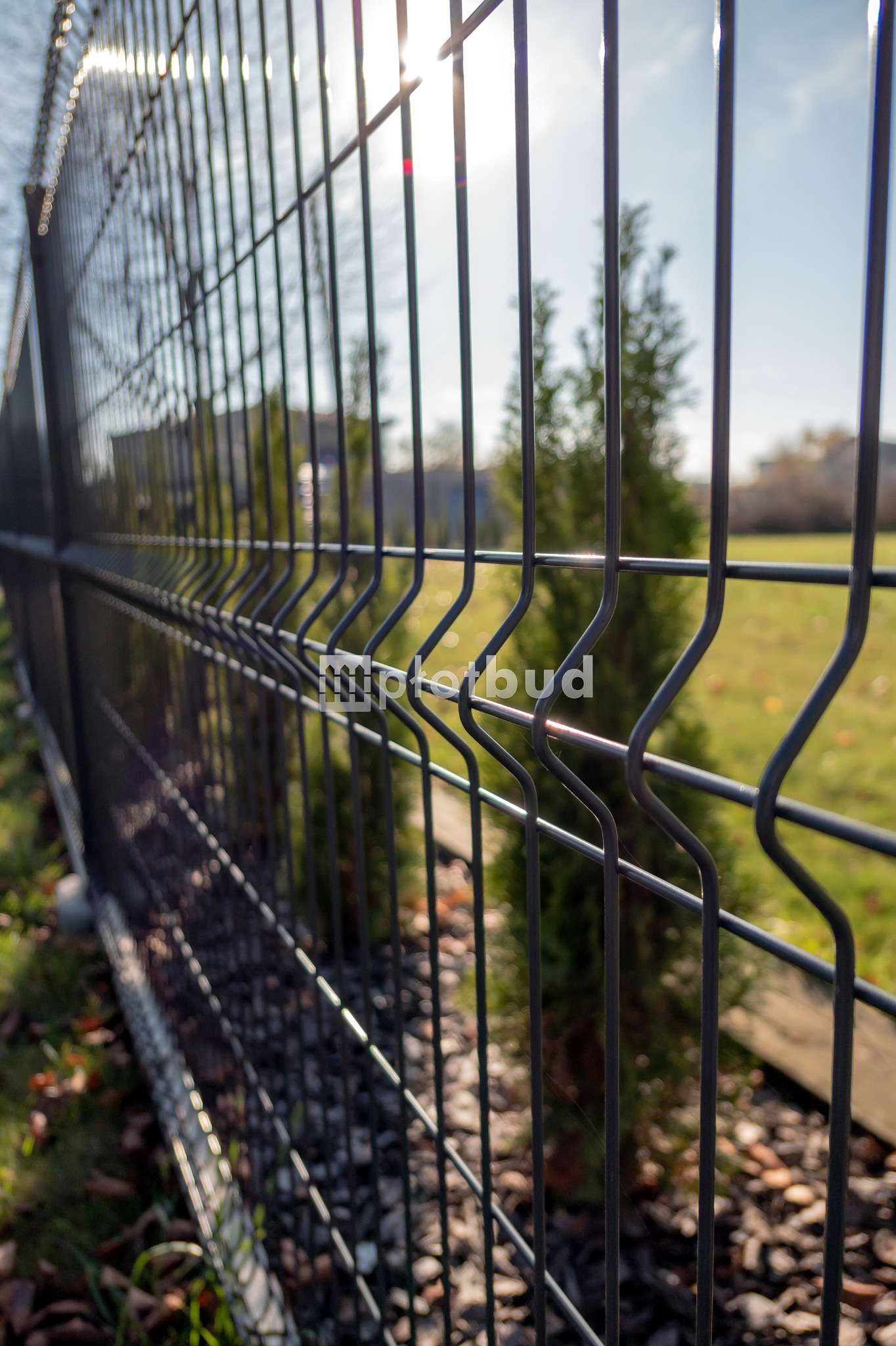 Ciemnoszare ogrodzenie panelowe z widocznym logotypem firmy na tle zielonego trawnika i iglaka, oświetlone promieniami słońca.