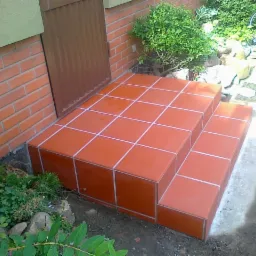 Wykończone płytkami ceramicznymi schody wejściowe do budynku, z widoczną fugą w kolorze białym i fragmentem elewacji z cegły.