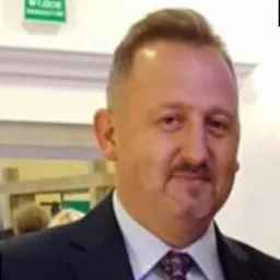 Portret mężczyzny w ciemnym garniturze i wzorzystym krawacie, z kr&oacute;tkimi włosami i wąsami, na rozmytym tle wnętrza z elementami dekoracyjnymi.