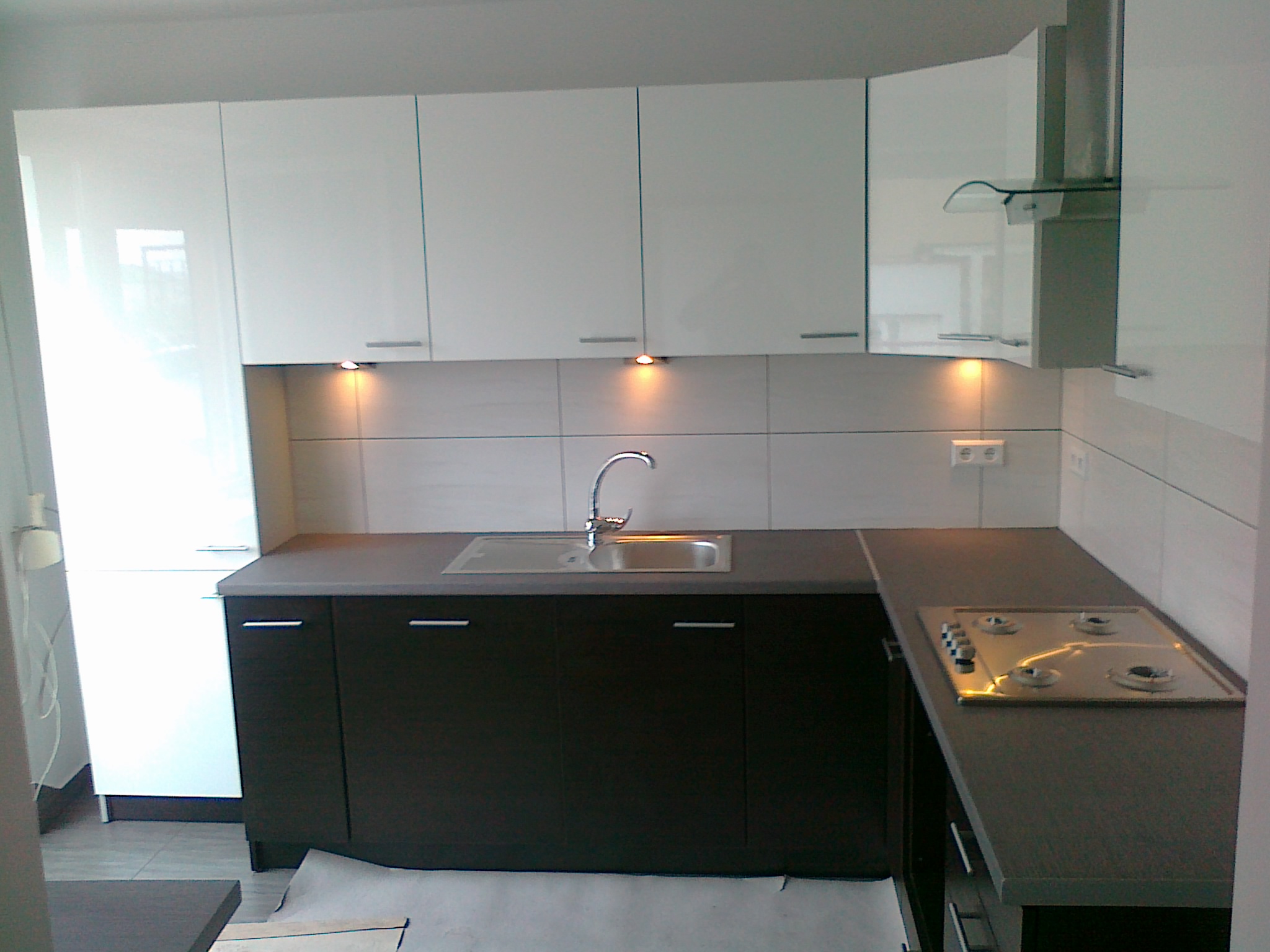 Nowoczesna kuchnia na wymiar z białymi szafkami wiszącymi i ciemnymi szafkami dolnymi, oświetleniem podszafkowym i płytą grzewczą na blacie.