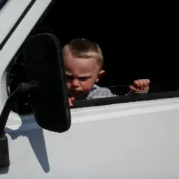 Mały chłopiec z poważną miną wygląda przez okno białego busa, trzymając się ramy okna, widoczne lusterko boczne.