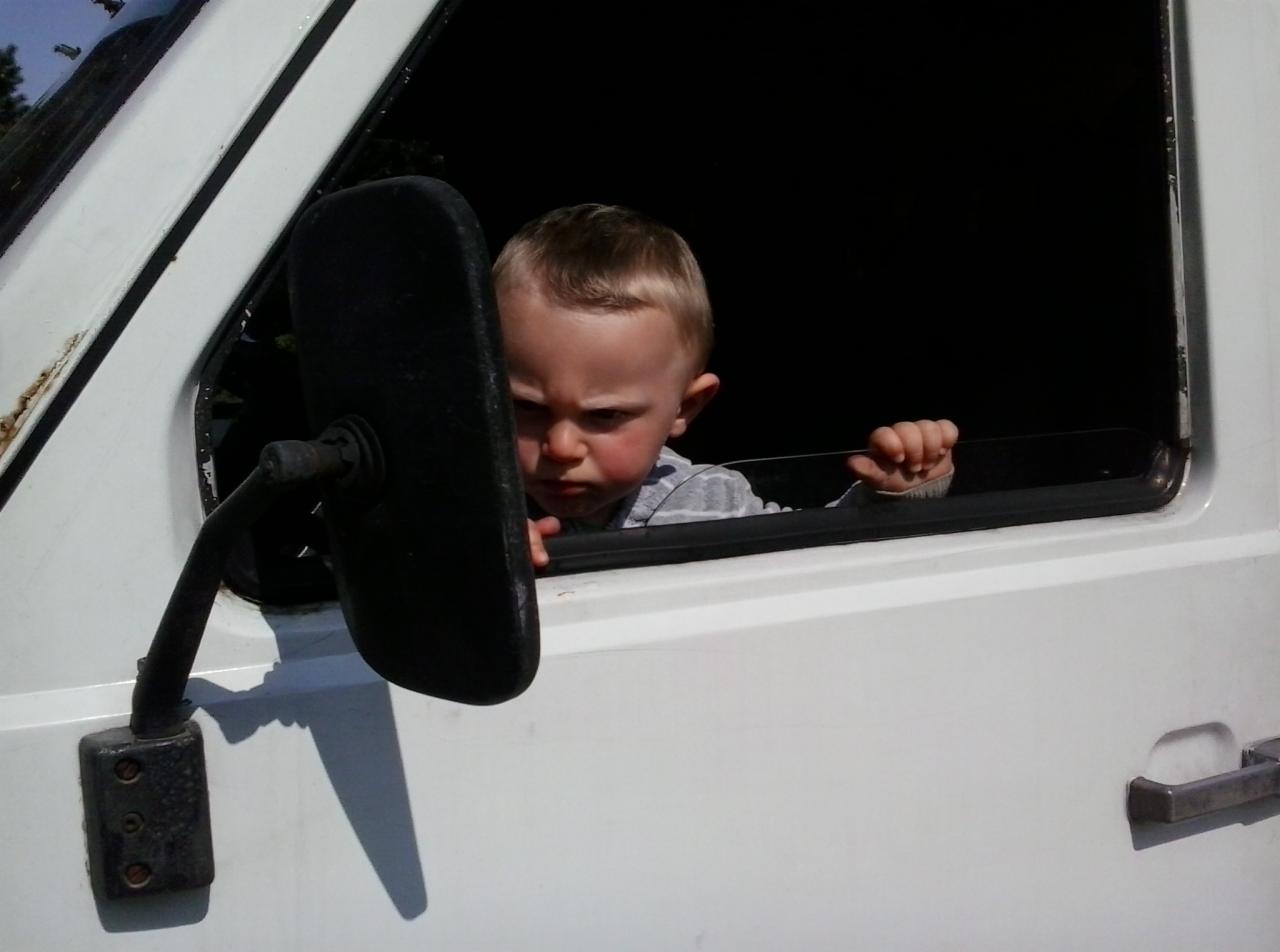Mały chłopiec z poważną miną wygląda przez okno białego busa, trzymając się ramy okna, widoczne lusterko boczne.