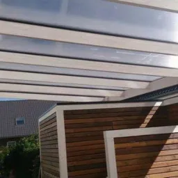 Zadaszenie tarasu z przezroczystym poliwęglanem i białymi belkami, widoczne drewniane panele ogrodowe i fragment dachu sąsiedniego budynku.