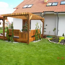 Drewniana pergola z ławkami i zadaszeniem, otoczona zielonym trawnikiem i ozdobnymi roślinami. Widoczny fragment domu z czerwonym dachem i okna dachowe.