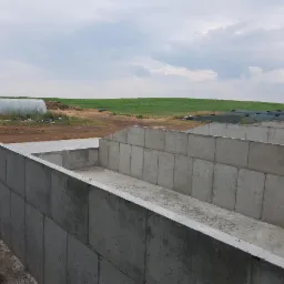 Betonowe silosy na polu uprawnym, widok z góry, na horyzoncie tunel foliowy i zielone wzgórza pod pochmurnym niebem.