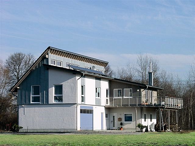 Dwukondygnacyjny dom z balkonem, z elementami prefabrykowanymi, wykończony w odcieniach szarości i bieli, z panelami słonecznymi na dachu i stosem drewna pod balkonem.