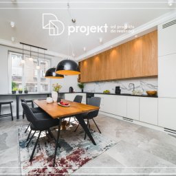 DD Projekt - Jasna kuchnia z drewnianymi i białymi szafkami, marmurowym blatem, drewnianym stołem z owocami, czarnymi lampami wiszącymi i dywanem w odcieniach szarości i czerwieni, z kotem leżącym pod stołem.