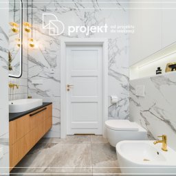 DD Projekt - Nowoczesna łazienka z marmurowymi płytkami, drewnianą szafką pod umywalką z czarnym blatem, złotą armaturą, wiszącą toaletą i bidetem, oraz białymi drzwiami.