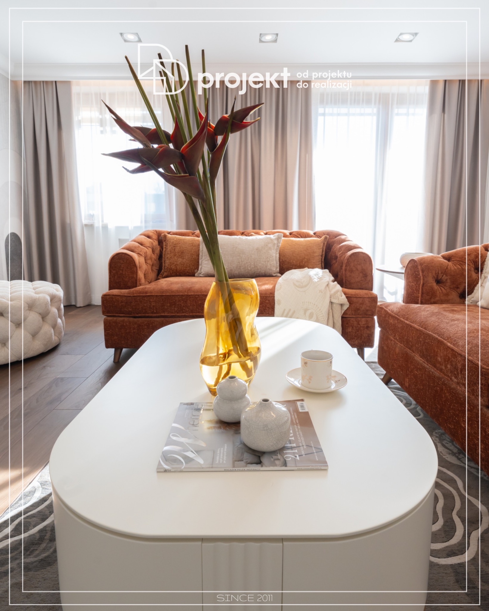 Salon z brązową sofą Chesterfield, białym pufem, owalnym stolikiem kawowym z magazynem wnętrzarskim, ceramicznymi figurkami i filiżanką, w tle okno z zasłonami.