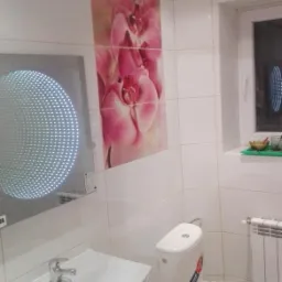 Biała łazienka z toaletą, umywalką z szafką, grzejnikiem i lustrem z efektem nieskończoności, ozdobiona płytkami z motywem różowych orchidei.