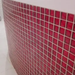 Zaokrąglona obudowa wanny wykończona drobną, czerwoną mozaiką z białymi fugami, widoczna bateria umywalkowa i fragment białej ściany z płytkami.