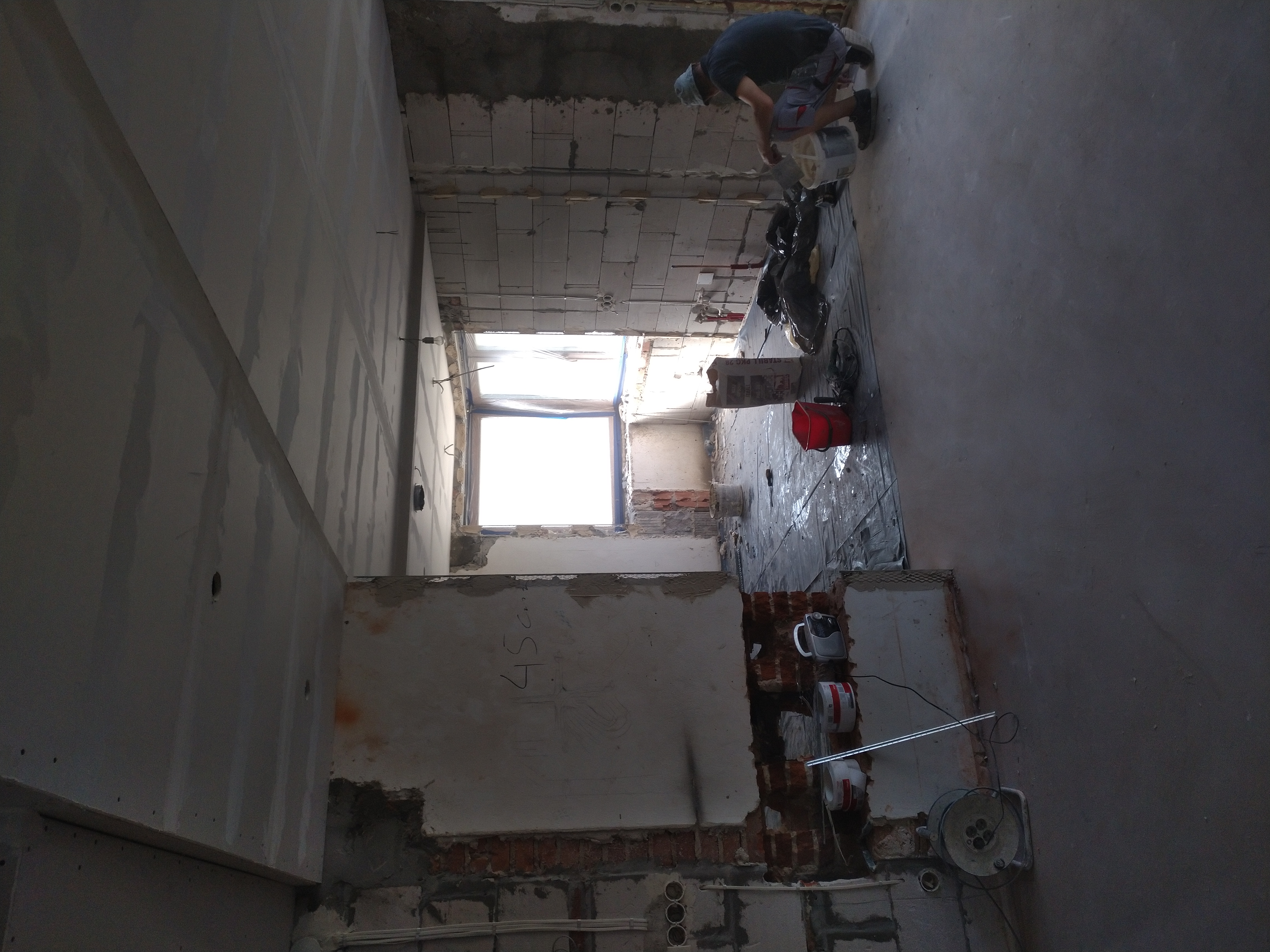 Prace przygotowawcze w trakcie remontu: pracownik miesza zaprawę, widoczne surowe ściany, instalacje elektryczne i okno w remontowanym wnętrzu.