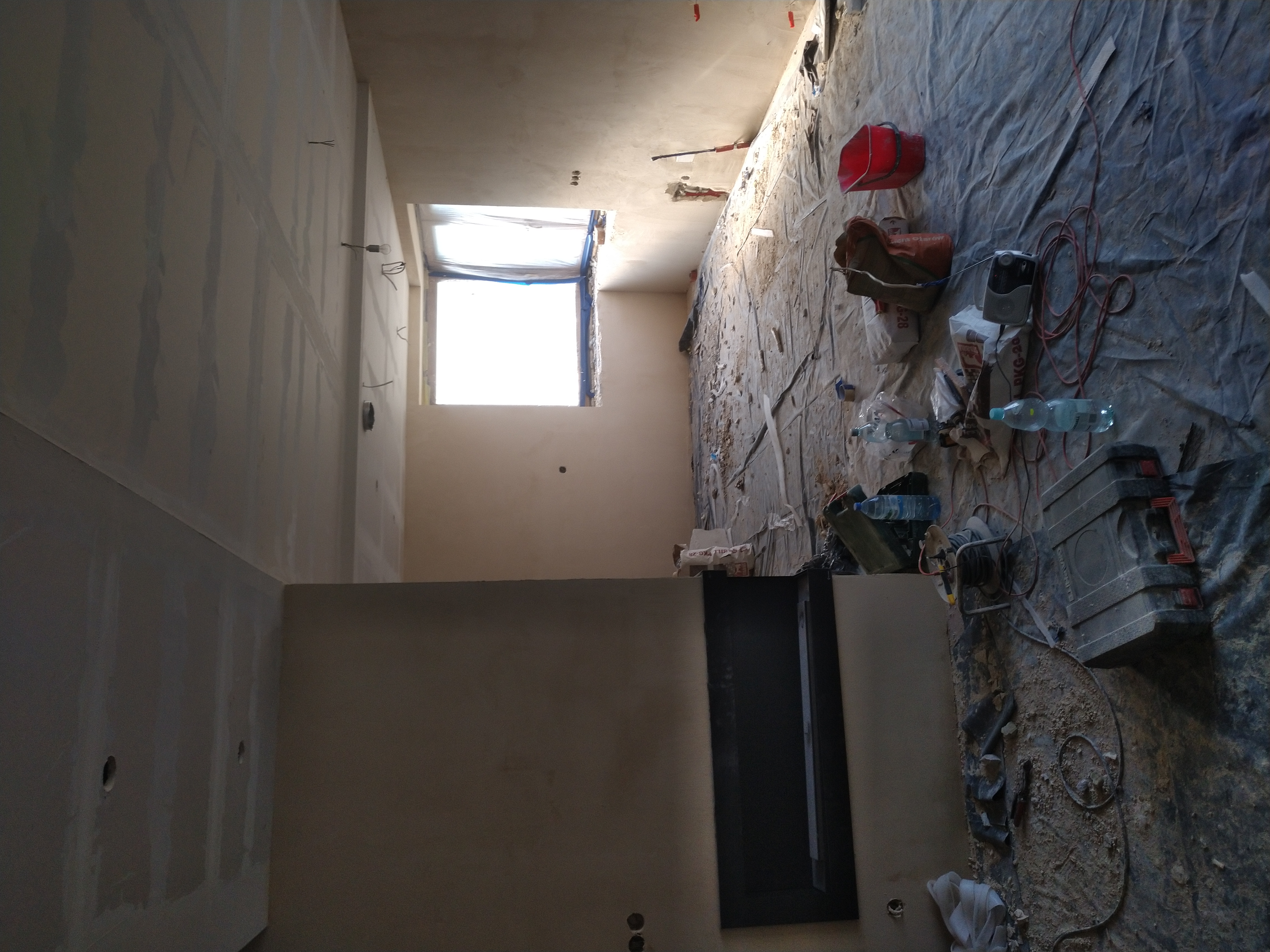 Wnętrze w trakcie prac wykończeniowych: ściany przygotowane pod gładzie, okno zabezpieczone folią, podłoga przykryta folią ochronną z widocznymi narzędziami i materiałami budowlanymi.