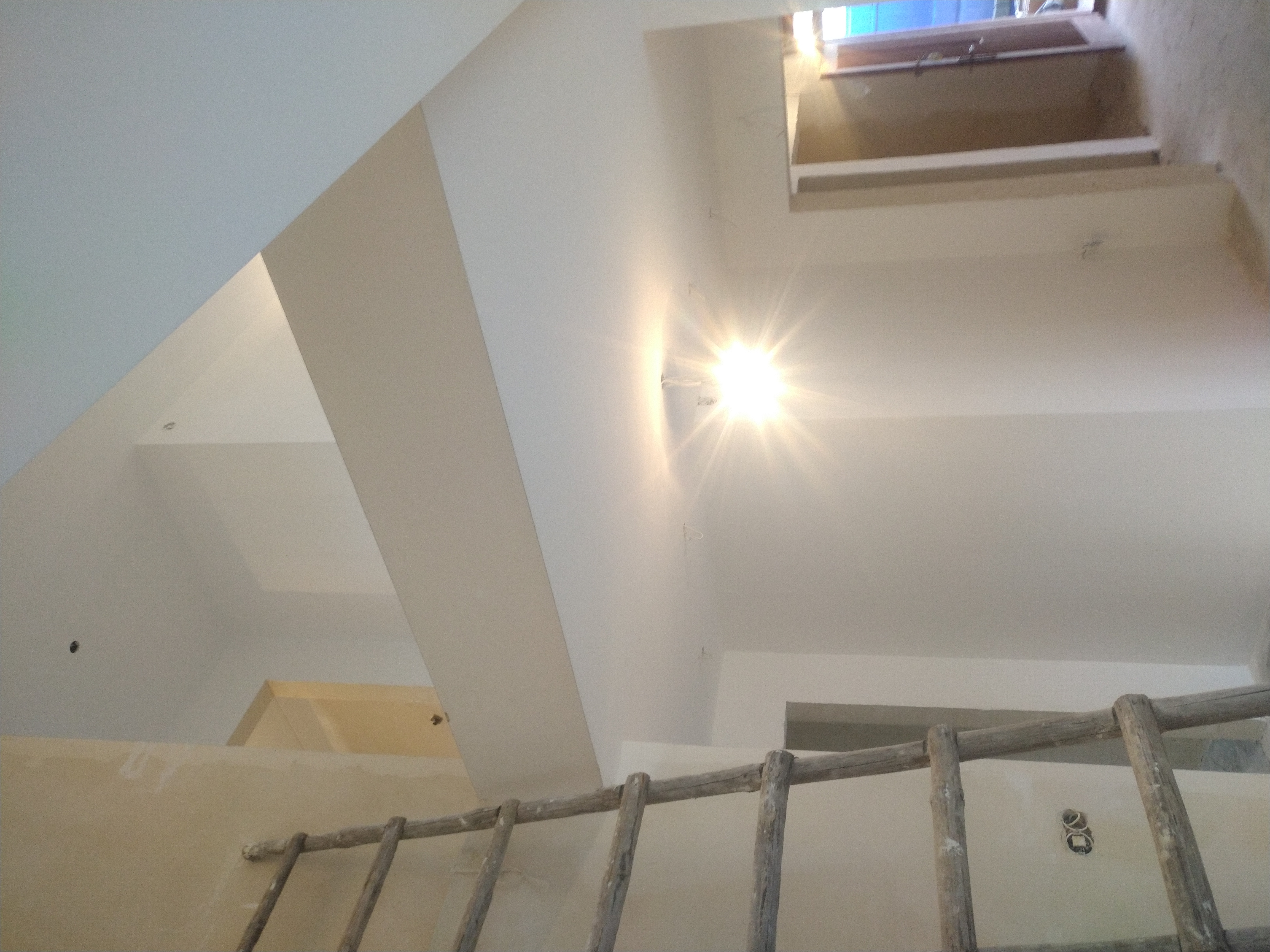 Wnętrze w trakcie prac wykończeniowych, widok z dołu na białe, gładkie ściany i sufit poddasza z oknem dachowym, fragment drewnianej drabiny i niedokończone punkty elektryczne.