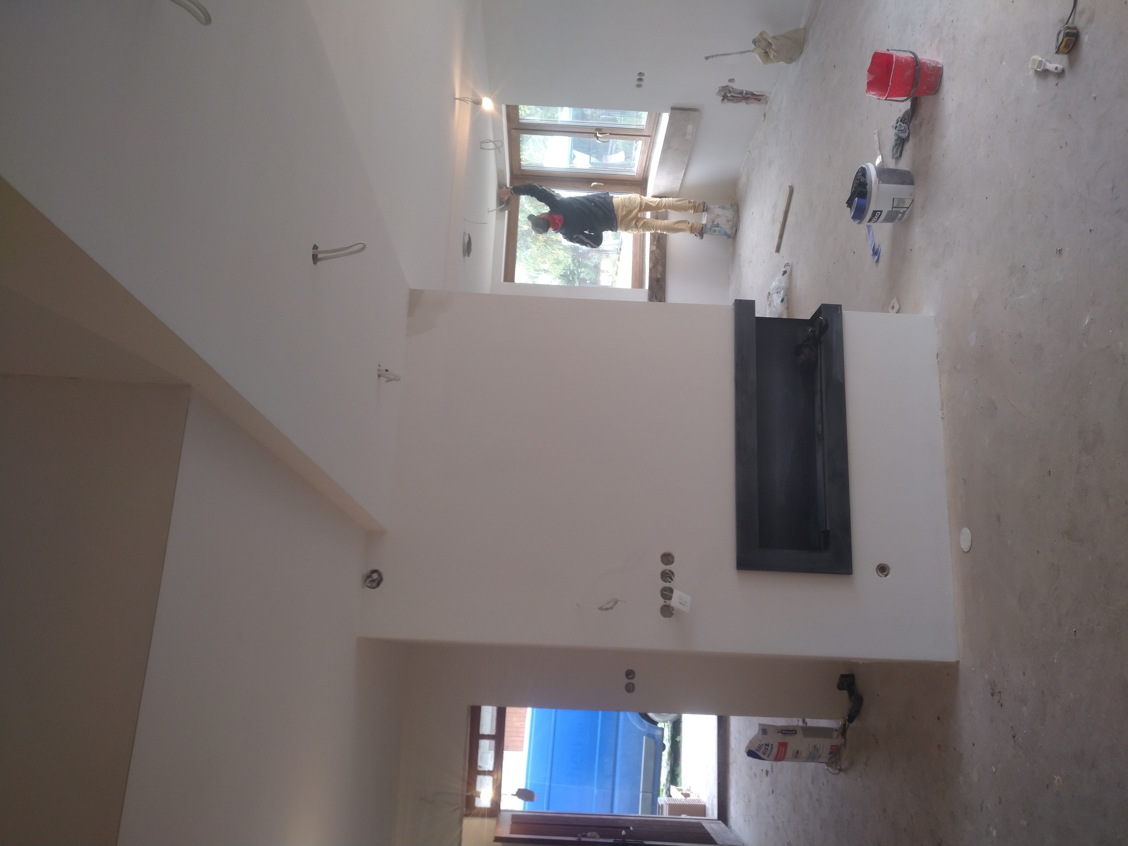 Pracownik w trakcie nakładania gładzi na suficie w nowym mieszkaniu, widoczne ślady po instalacjach elektrycznych, okno i fragment ściany z kominkiem.