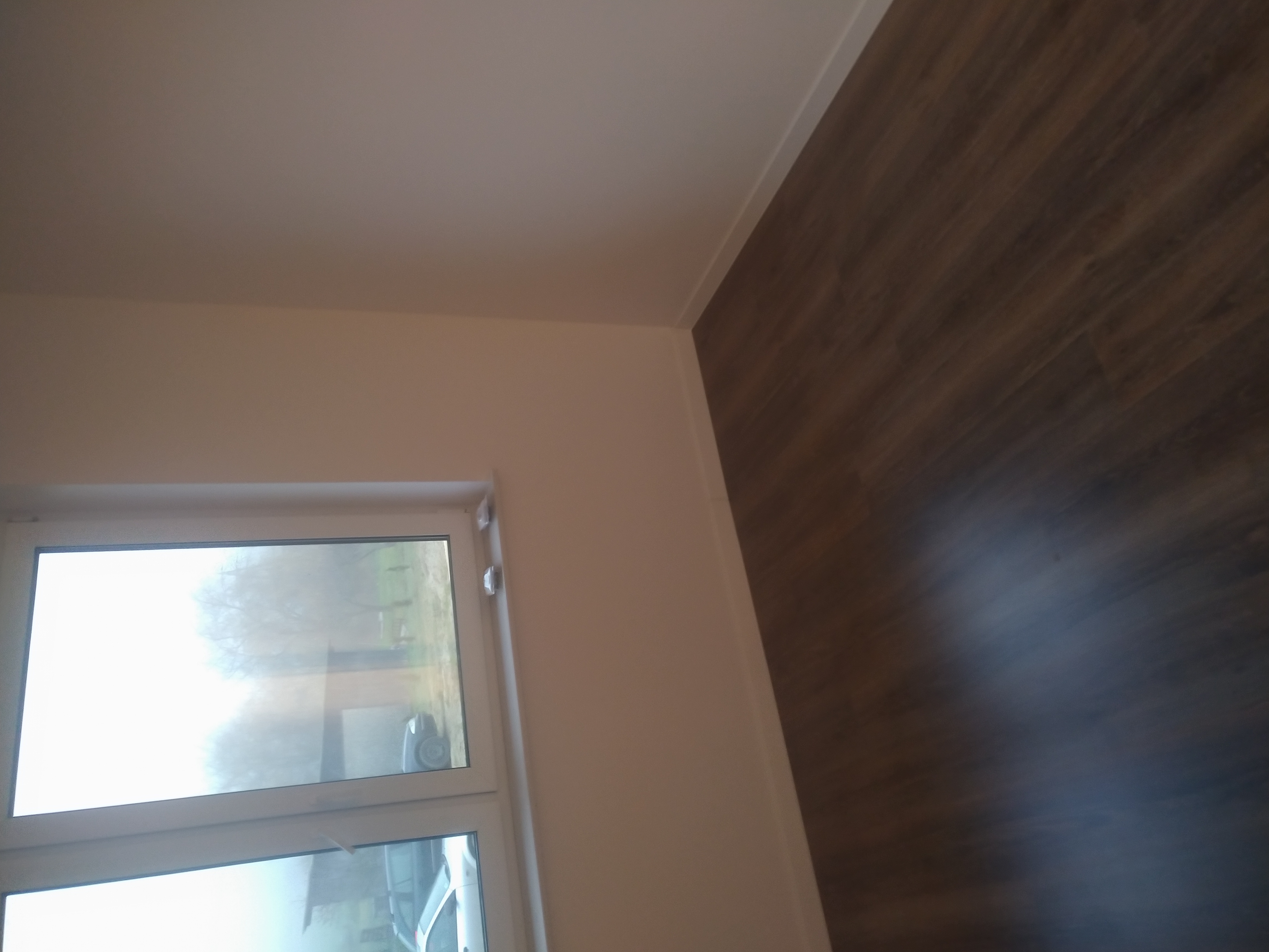 Fragment pokoju po remoncie: świeżo pomalowana ściana w odcieniach beżu, panelowa podłoga imitująca drewno, białe listwy przypodłogowe i okno z widokiem na zewnątrz.