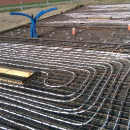 Przygotowanie podłoża pod wylewkę betonową z ułożonym ogrzewaniem podłogowym, widoczna siatka zbrojeniowa i rury instalacji grzewczej w białym kolorze, na zewnątrz budynku.