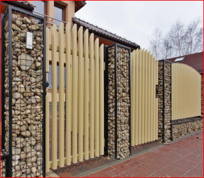 Nowoczesne ogrodzenie gabionowe z beżowymi pionowymi panelami, wypełnione kamieniami o różnej wielkości, z czarną metalową konstrukcją, na tle ceglanej ścieżki i budynku.