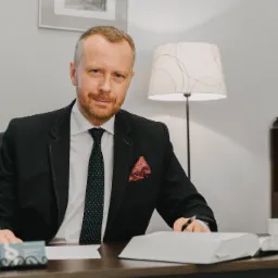 Portret mężczyzny w garniturze siedzącego za biurkiem z otwartą księgą, wizytówką i przyborami do pisania, w tle lampa i regał z książkami.