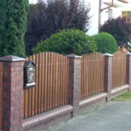 Nowoczesne ogrodzenie z drewnianych sztachet i ceglanych słupków, zwieńczonych ciemnymi daszkami, z zamontowaną skrzynką pocztową, na tle zieleni i domu.