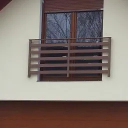 Brązowa, pozioma balustrada balkonowa zamontowana na elewacji budynku z oknem i roletą zewnętrzną, widok z dołu na fragment budynku z garażem.