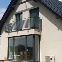 Nowoczesny dom z szarą, metalową balustradą balkonową o poziomym układzie szczebli, widoczny z zewnątrz, z fragmentem placu budowy w tle.