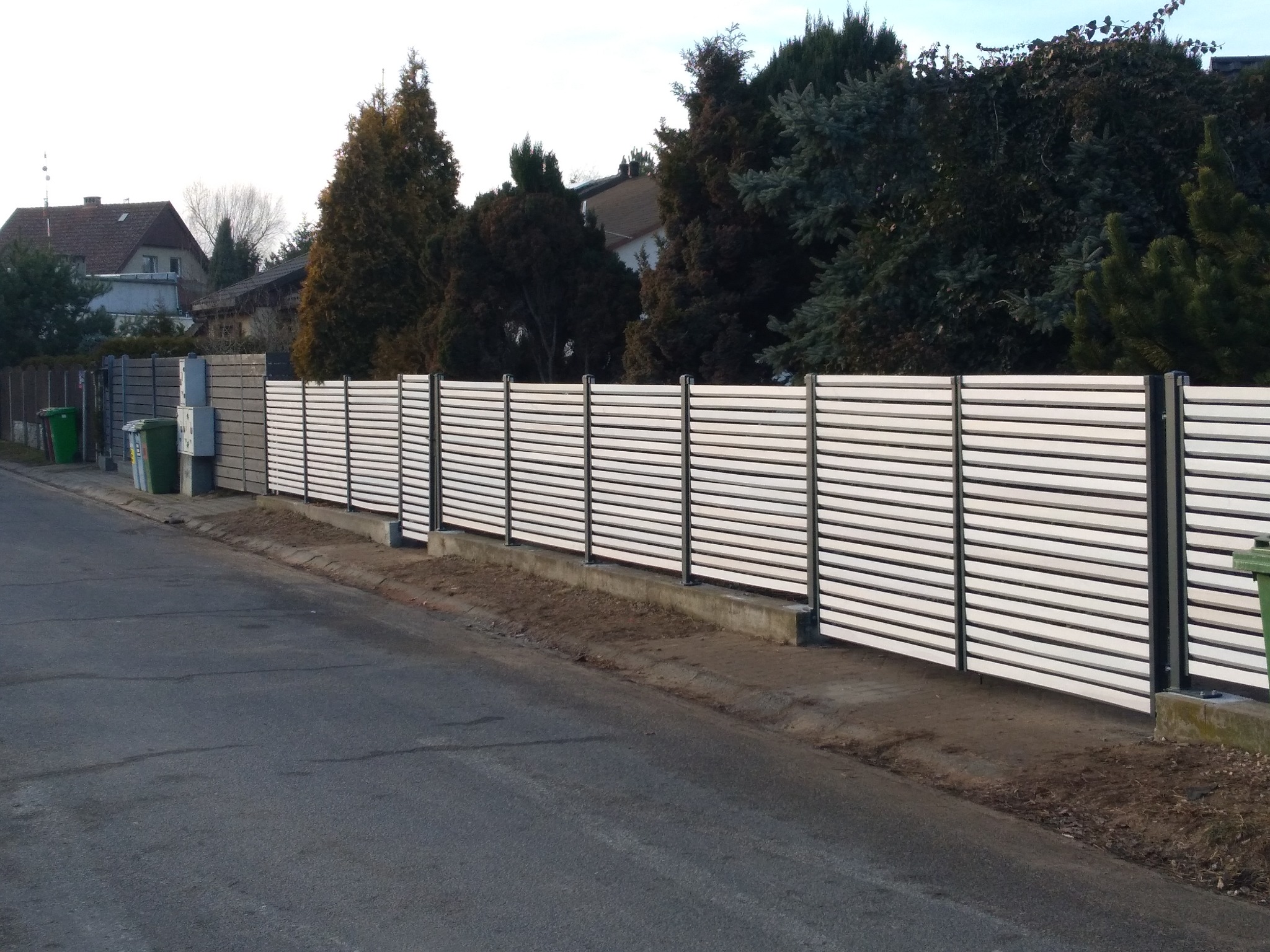 Nowoczesne ogrodzenie ażurowe z białych, poziomych desek zamontowanych na czarnych słupkach, oddzielające posesję od ulicy.