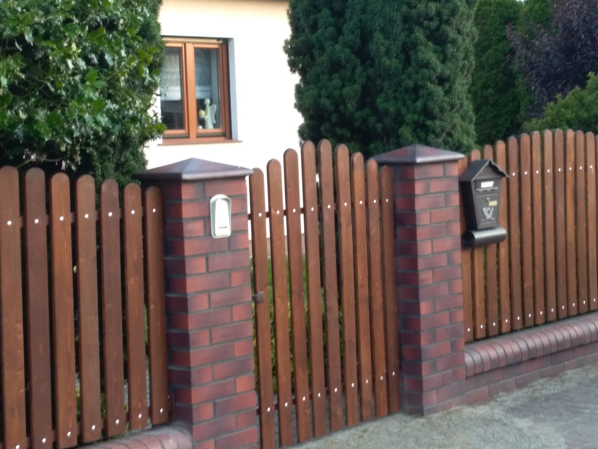 Brunatne ogrodzenie sztachetowe z ceglanymi słupkami i skrzynką na listy, widok na dom z oknem i zielenią.