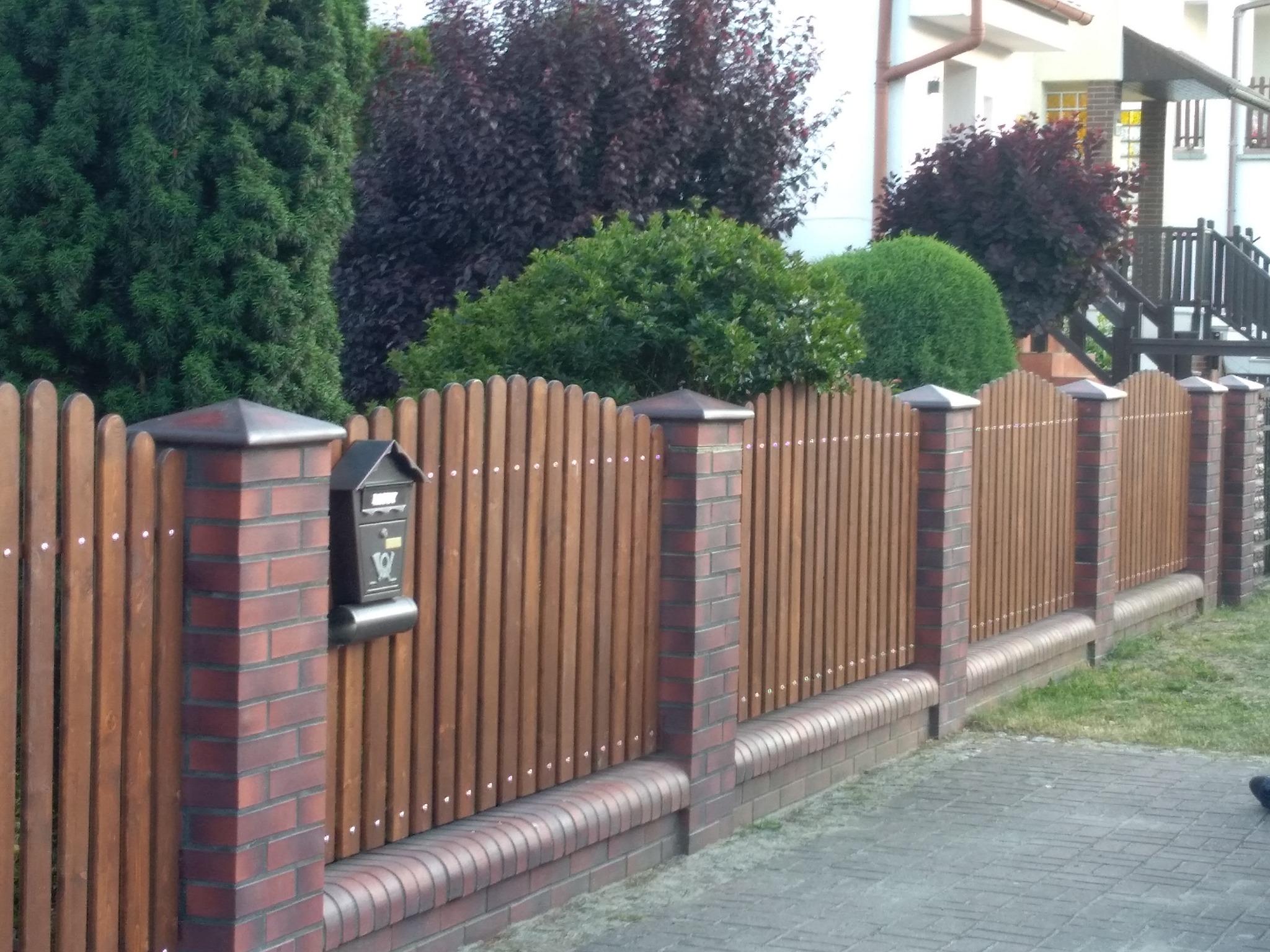 Nowoczesne ogrodzenie z drewnianych sztachet i ceglanych słupków, zwieńczonych ciemnymi daszkami, z zamontowaną skrzynką pocztową, na tle zieleni i domu.