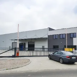 Nowoczesny budynek magazynowy z bramą wjazdową i szarym sedanem zaparkowanym obok, na pierwszym planie kostka brukowa i asfaltowa droga.