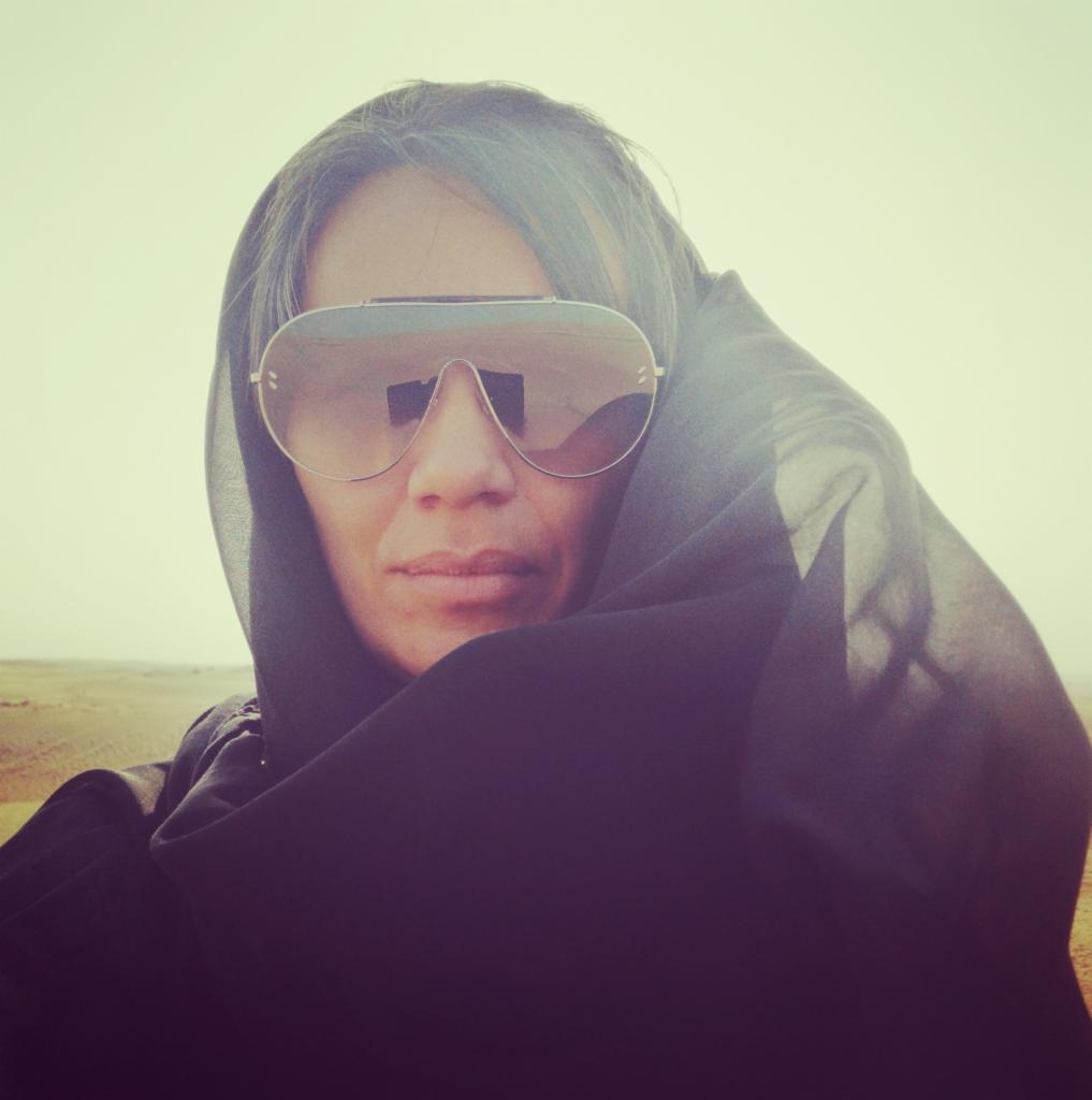 Portret kobiety w okularach lotniczych i czarnym nakryciu głowy na tle pustynnego krajobrazu w ciepłych tonacjach.