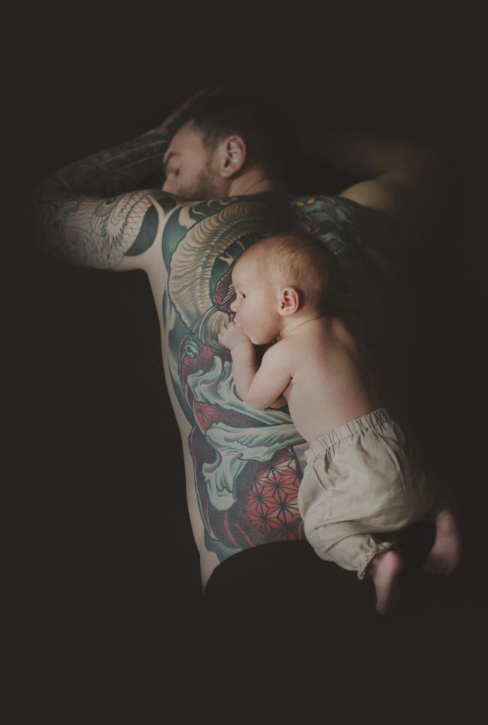 Niemowlę w beżowych spodniach leży na wytatuowanych plecach mężczyzny, twarzą w stronę obiektywu, w ciemnym otoczeniu.