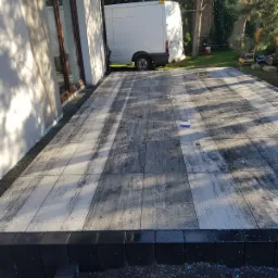 Świeżo ułożona, wilgotna nawierzchnia z szarych płyt z efektem betonu, obramowana czarną kostką, z widoczną białą furgonetką w tle i szczotką do zamiatania na powierzchni.