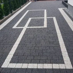 Nowoczesny podjazd z szarej kostki z białymi, geometrycznymi wzorami, obrzeżami i pasem żwiru przy ścianie budynku.