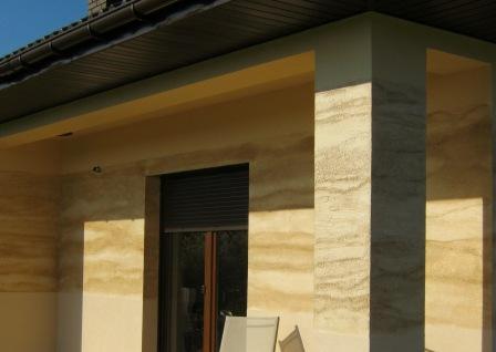 Elewacja budynku z dekoracyjnym tynkiem imitującym trawertyn, widoczny fragment okna z żaluzjami zewnętrznymi oraz kolumna w tym samym stylu, zarys dachu w tle.
