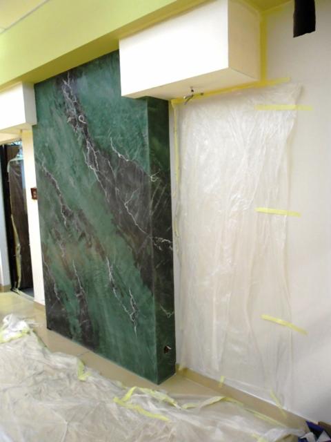 Ściana z efektem dekoracyjnym imitującym zielony marmur, częściowo zakryta folią ochronną przyklejoną taśmą malarską, podłoga zabezpieczona folią, widoczne przygotowania do prac wykończeniowych.