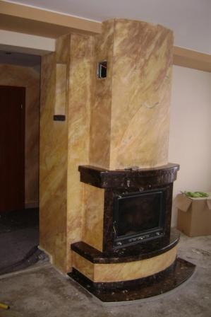 Kominek z obudową w tynku dekoracyjnym imitującym trawertyn, z czarnym marmurowym wykończeniem paleniska, widoczne niedokończone prace wykończeniowe.