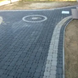 Ułożona nawierzchnia z kostki brukowej w odcieniach szarości z dekoracyjnym okręgiem i jasnoszarym obramowaniem, widoczny fragment wózka budowlanego oraz schody wejściowe do budynku.