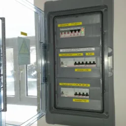 Otwarta szara skrzynka rozdzielcza z widocznymi bezpiecznikami i oznaczeniami gniazd 1 i 3 fazowych, zamontowana na białej ścianie, obok okna z naklejką ostrzegawczą.