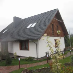 Nowoczesny dom jednorodzinny z ciemną dachówką, oknami dachowymi i drewnianą elewacją szczytową, z zadaszonym tarasem i zagospodarowanym ogrodem z oświetleniem ścieżki.