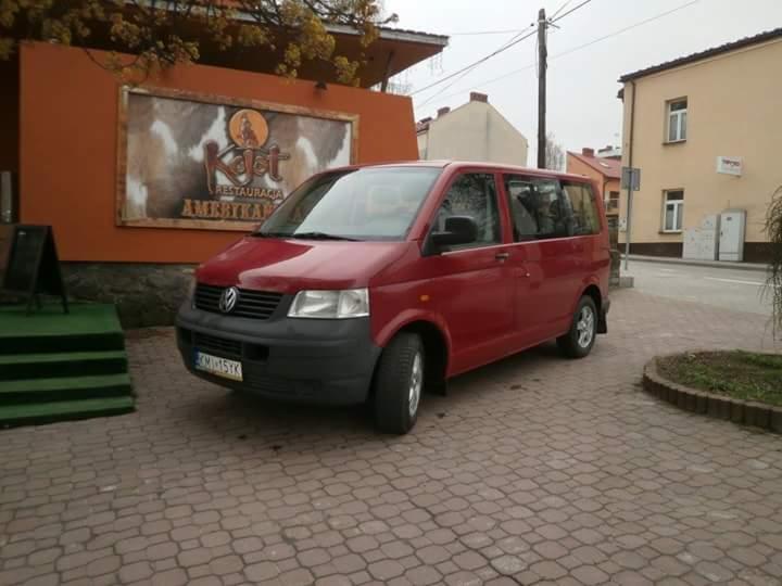 Czerwony Volkswagen Transporter zaparkowany na brukowanym chodniku przed budynkiem z szyldem restauracji 'Kojot'.