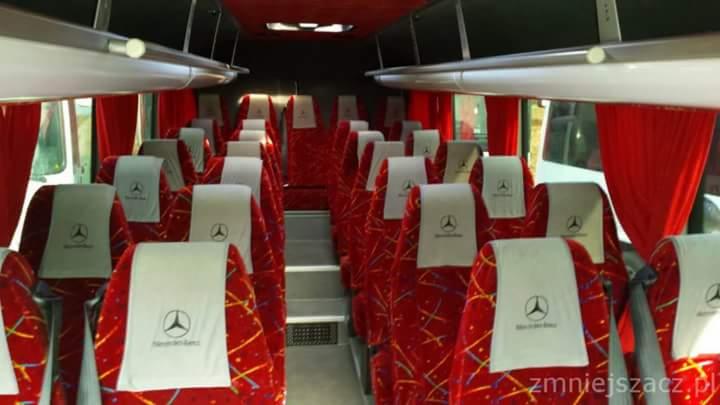 Wnętrze autobusu z czerwonymi siedzeniami z białymi zagłówkami z logo Mercedes-Benz, widok z przodu, czerwone zasłony i szare schodki na środku przejścia.