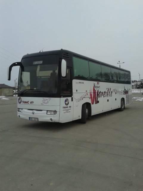 Biały autokar z czarnym dachem, z logo firmy Trans RD na boku, zaparkowany na betonowym placu w pochmurny dzień.