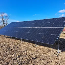 Rząd paneli słonecznych na metalowej konstrukcji, umieszczonych na polu z resztkami pożniwnymi, błękitne niebo z chmurami w tle.